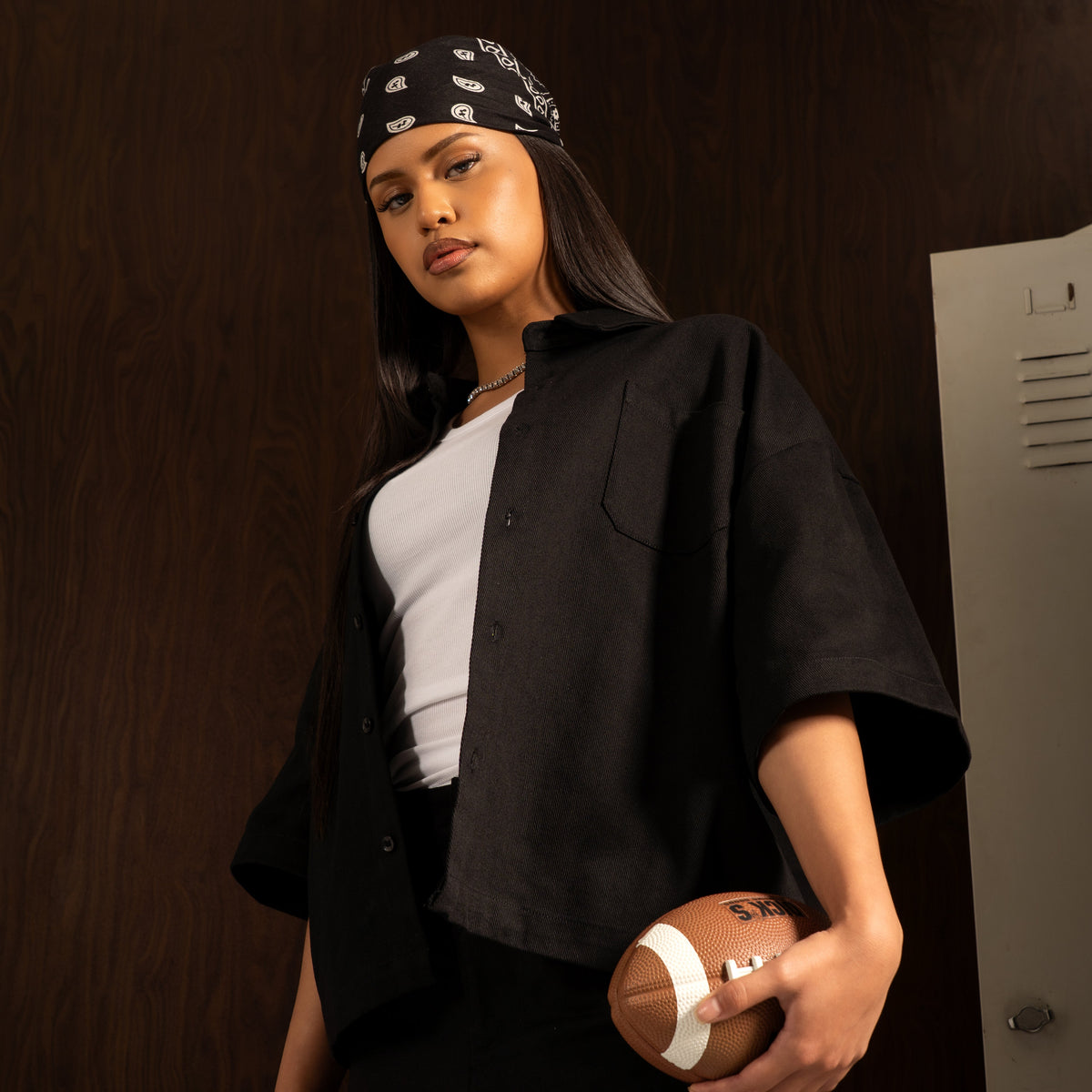 A woman stands in a locker room holding a football, wearing a black oversized button-up shirt over a white tank top, black pants with a white graphic, and a black bandana with white logos.